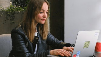 A imagem mostra uma mulher branca de cabelos castanhos claros, longos e lisos, usando uma blusa branca e uma jaqueta de couro preta. Ela está em um escritório, sentada em uma cadeira e olhando seriamente para um computador aberto. Ela acaba de receber uma cobrança judicial da Capes ou do CNPq.