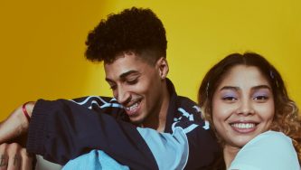 Um casal jovem e sorridente posa juntos contra um fundo amarelo vibrante. O homem, pardo, com cabelo encaracolado curto e uma camiseta escura, descansa sua cabeça levemente sobre o ombro da mulher. A mulher, exibindo cachos soltos e uma blusa de cor clara, Ambos exalam uma sensação de conforto e afeto compartilhado, sugerindo uma conexão próxima e um espírito jovial.