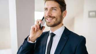 A imagem mostra um homem branco, de cabelos castanhos e curtos e olhos azuis. Ele usa uma blusa branca, um terno azul escuro e uma gravata também azul. Ele olha para a frente sorrindo, com uma das mãos segurando o celular no ouvido.
