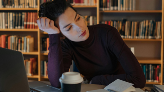 A imagem mostra uma mulher jovem e branca, de cabelos pretos e presos em um rabo, usando uma blusa de frio azul. Ela está em uma biblioteca (tem uma estante cheia de livros atrás) e sentada em uma cadeira, com um laptop aberto em uma mesa a sua frente. Ela está com a cabeça apoiada em uma das mãos enquanto olha para o lado com um olhar desolador pensando sobre a sua desistência de bolsa de estudos no exterior.