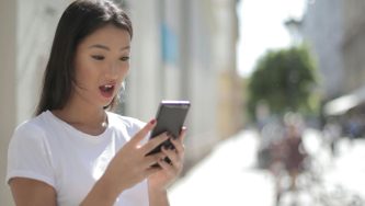 A imagem mostra uma mulher em uma rua. Ela é jovem, branca, tem cabelos lisos, pretos e soltos. Ela usa uma blusa branca de mangas curtas. Ela está segurando um celular com as duas mãos e olhando para ele com uma cara de espanto.