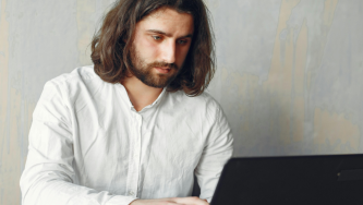A imagem mostra um homem brance de barba e cabelos lisos e soltos até o ombro, usando uma blusa social branca, de manga longa. Ele está em um escritório de paredes cinzas, sentado em uma cadeira e com as mãos em um computador. Ele olha atentamente para o computador enquanto estuda questões relativas a dívida trabalhista de ONG.