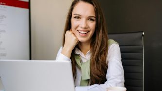 A imagem mostra uma mulher branca de cabelos marrons, longos, lisos e soltos. Ela está em um quarta, sentada em uma cadeira e com os cotovelos apoiados em uma mesa, com um computador aberto no Domicílio Judicial Eletrônico. A mão direita está apoiada no queixo e ela sorri para a câmera.