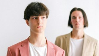 A imagem mostra dois homens representando o empreendedorismo LGBT+ O que está mais ao fundo é branco, cabelos castanhos, lisos e soltos em formato chanel, usa uma camisa branca e um paletó bege. O homem da frente é branco, tem cabelos curtos e castanhos, usa uma camisa branca e um paletó rosa.