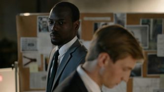 A imagem mostra dois homens em um ambiente de trabalho. O homem em primeiro plano, à direita, está levemente desfocado, enquanto o homem ao fundo, à esquerda, usa terno e gravata e observa a cena com expressão atenta. Ele o observa pensando em como garantir extensão do regime jurídico.