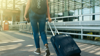 A imagem mostra uma mulher branca de costas caminhando pela área externa de um aeroporto. Ela usa uma mochila nas costas, uma calça jeans e um tênis enquanto puxa uma mala de rodinhas. Ela está buscando informacões sobre extravio de bagagem após sair de um voo.