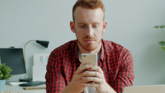 A imagem mostra um homem branco de cabelos ruivos e barba ruiva, usando uma blusa branca e uma camiseta xadrez vermelha e preta por cima. Ele observa o celular com expressão preocupada, buscando dados de golpe virtual do qual foi vítima. Ele está sozinho e em um escritório bem iluminado.