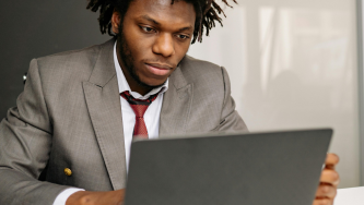 A imagem mostra um homem negro de cabelos marrons em dreads. Ele usa uma blusa branca e um terno cinza com gravata vermelha. Ele está sentado diante de um computador aberto enquanto lê sobre governança de dados no Terceiro Setor. Ele olha para a tela com expressão concentrada, apoiando uma das mãos próxima ao computador. Ao fundo, há um ambiente interno desfocado, com tons claros.