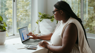 A imagem mostra uma mulher negra de cabelos pretos, lisos e longos, trabalhando com um computador, sentada em frente a uma mesa próxima a uma janela, com boa iluminação natural. Na mesa há ainda uma calculadora e um bloco de notas e, ao fundo, algumas plantas. A mulher calcula se tem direito a hora extra no home office.