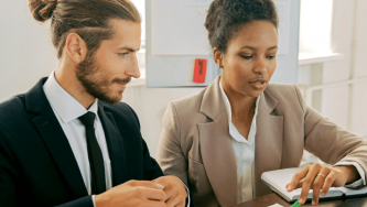 A imagem mostra dois profissionais em um ambiente corporativo, sentados lado a lado em uma mesa de trabalho. O da esquerda é um homem branco de cabelos castanhos, longos, presos em um coque, usando um terno preto e gravata. A da direita é uma mulher negra de cabelos crespos, longos e presos, usando uma blusa social branca e um blazer marrom. Eles analisam documentos e anotações a respeito de impostos sobre herança.