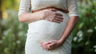 A imagem mostra uma a barriga de uma mulher grávida. Ela está em um ambiente aberto e com jardins, usa um vestido branco e coloca as mãos na barriga. Essa mulher vai poder desfrutar da nova lei da Licença-Maternidade Estendida.
