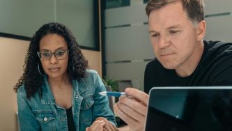 A imagem mostra uma mulher de cabelos enrolados e soltos, óculos de grau, blusa preta e casaco jeans. Ao seu lado esquerdo está um homem de cabelos curtos com uma caneta na mão. Ambos estçao sentados a frente de uma mesa e olham fixamente para um docimento, posicionado acima da mesa.