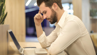 Um homem branco de barba curta e camisa social branca está sentado em uma cadeira a frente de uma mesa de madeira. Ele está inclinado para frente, com o cotovelo apoiado na mesa e a mão na testa. À sua frente há um notebook aberto onde ele lê sobre metas abusivas no trabalho. Ao lado há um celular e uma xícara de café.