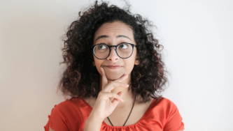 Uma mulher com cabelo cacheado e volumoso aparece em frente a um fundo claro, usando uma blusa laranja e óculos de armação escura. Ela está com a mão apoiada no queixo, olhando para cima, com expressão pensativa sobre negociar dívida com Capes ou CNPq.