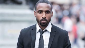 A imagem mostra um homem negro de cabelos curtos e cavanhaque com expressão séria, vestindo terno preto, camisa branca e gravata, caminhando em um ambiente urbano. A postura firme e o olhar direto transmitem segurança e foco, elementos que remetem à importância da atuação profissional em temas complexos, como a nulidade do PAD (Processo Administrativo Disciplinar).