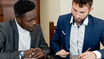 A imagem mostra dois homens sentados à mesa, em um ambiente interno. O homem da esquerda é negro, tem caeblos crespos, pretos e curtos, veste uma blusa branca e um paletó preto. O homem da direita é branco e tem cabelos lisos, marrons e curtos. Ele usa uma blusa branca e um paletó azul escuro. Eles estão inclinados sobre a mesa e o homem da direita assina um testamento.