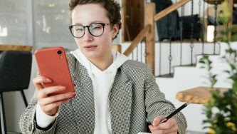Uma mulher branca de cabelos ruivos e presos em uma trança está sentada à mesa em um ambiente interno, segurando um celular vermelho com a mão esquerda e uma caneta com a mão direita, como se estivesse lendo algo na tela enquanto faz anotações sobre como pejotização no Terceiro Setor. Ela usa óculos de armação escura, blusa clara com capuz e um blazer cinza.