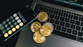 A imagem mostra uma mesa onde vemos um computador aberto, um celular no aplicativo de calculadora e moedas prateadas e douradas representando criptomoedas.