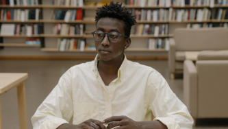 A imagem mostra um homem jovem, negro de cabelos pretos e curtos. Ele usa um óculos de grau e uma blusa social de tom creme. Ele está em uma biblioteca, com uma estante de livros com capas de diversas cores ao fundo e algumas mesas ao redor. Ele está sentado em uma cadeira, com os braçoes recostados em uma mesa e uma expressão pensativa sobre como realizar prestação de contas após aprovação da novação