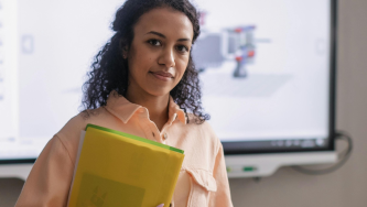 A imagem mostra uma mulher de pele negra com cabelos cacheados presos para trás. Ela está em pé, olhando para frente, segurando pastas coloridas sobre progressão de carreira de professor universitário. Ela veste uma camisa de tom pêssego e ao fundo vemos uma tela, onde há um desenho técnico.
