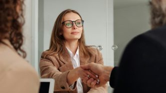 A imagem mostra as costas de duas pessoas nos cantos direito e esquerdo da foto e, ao fundo, uma mulher a frente deles. Ela é uma mulher branca de cabelos lisos, castanhos e soltos, com óculos de grau, vestindo uma blusa branca e um paletó bege. Ela está olhando para a pessoa da direita com um olhar sério e aperta a sua mão. Entre eles está uma mesa, o ambiente parece um escritório.