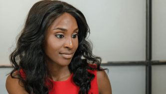 A imagem mostra uma mulher negra de cabelos pretos e ondulados até a altura do ombro. Ela usa uma blusa vermelha e está sentada em uma sala de escritório bem iluminada. Ela olha para a frente como se estivesse olhando para um computador em busca de informações sobre reintegração de candidata eliminada em concurso.