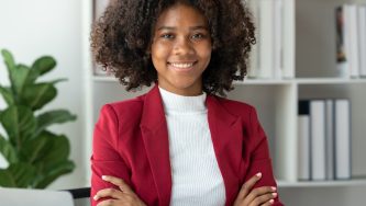 A imagem mostra uma mulher jovem, negra, de cabelos crespos, soltos até a altura do ombro. Ela está com uma blusa branca e um blazer de cor vemelho escura. Seus braços estão cruzados e ela olha para a câmera com um sorriso confiante. Ela está em um ambiente fechado, com uma biblioteca ao fundo e um vaso de plantas.