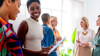 A imagem mostra uma sala com várias mulheres em uma sala fechada de paredes brancas e iluminada por uma grande janela. Ao fundo, quatro mulheres com roupas sociais conversando. A frente duas mulheres de pele negra e roupas sociais conversam. A que está mais à direita tem um papel nas mãos. Elas olham uma para a outra e sorriem.
