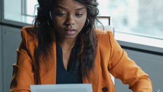 A imagem mostra uma mulher negra de cabelos grandes, ondulados e soltos, usando uma blusa preta e um blazer da cor mostarda. Ela está em um escritório, com uma janela com vista para a cidade ao fundo, sentada em uma cadeira e com os olhos fixos para a tela de um computador que está aberto à sua frente.
