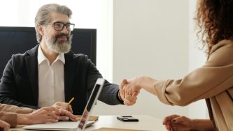 A imagem mostra um homem branco de cabelos e barba grisalhos. Ele usa um óculos de grau, blusa social branca e um paletó preto por cima. Ele está em um escritório, sentado à mesa de reunião com outras pessoas, Ele olha com um sorriso para uma mulher de cabelos enrolados e soltos que está de costas para quem olhando para a imagem. Os dois estão apertando as mãos.