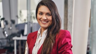 A imagem mostra uma mulher branca de cabelos lisos, pretos e soltos. Ela usa uma blusa branca e um casaco vermelho escuro e segura uma pasta com as mãos. Ela está em um ambiente empresarial iluminada e sorri para a câmera.